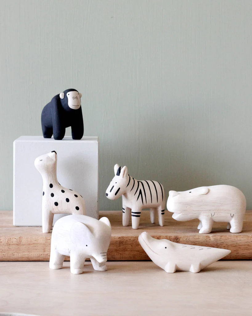 A collection of six hand-carved wooden animal figurines, crafted from handcrafted Albizia wood, includes a black monkey, a white giraffe with black spots, a white zebra with black stripes, a white rhinoceros, a white elephant, and a white crocodile. They're arranged on a wooden surface with a pale green background.