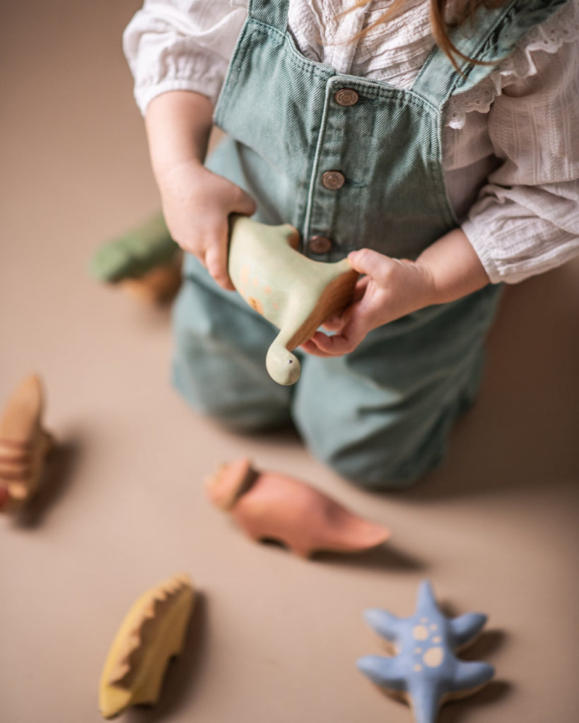 A toddler in overalls and a white shirt plays with a Handmade Wooden T-rex Dinosaur, holding a green watering can and pouring out invisible water. The toys, painted with non-toxic paint, include a sun