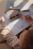A child in patterned clothing colors in My Coloring Book with crayons, placed beside a tin box on a white table, illuminated by natural light.
