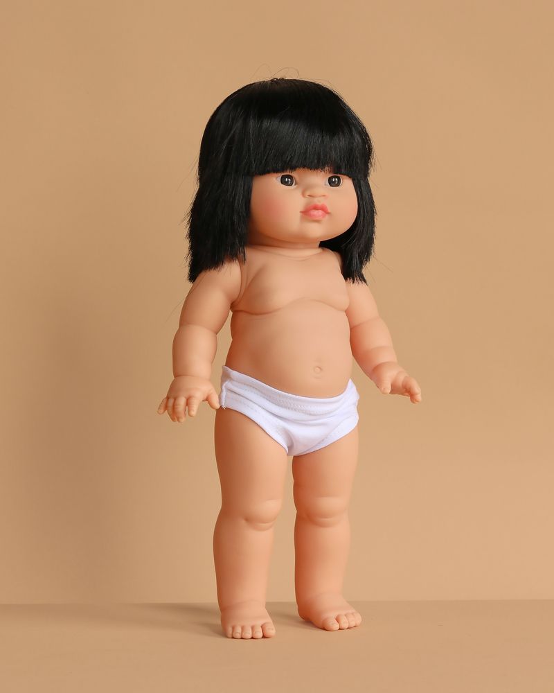 The Minikane Standing Baby Doll (14") - Jade features straight black hair with bangs and comes dressed in white underwear. This anatomically correct doll, made in Spain, stands upright against a plain beige background. Clothing sold separately.