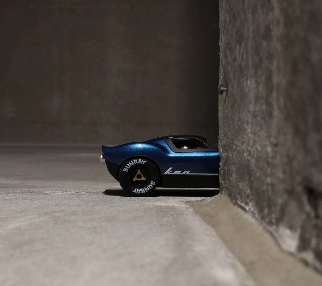 The Playforever MINI KEN CASH Blue, featuring “ken” on the side and “SUNRAY” tires, is pressed against a concrete wall in a dimly lit, empty room evoking the spirit of 1960s endurance racing.