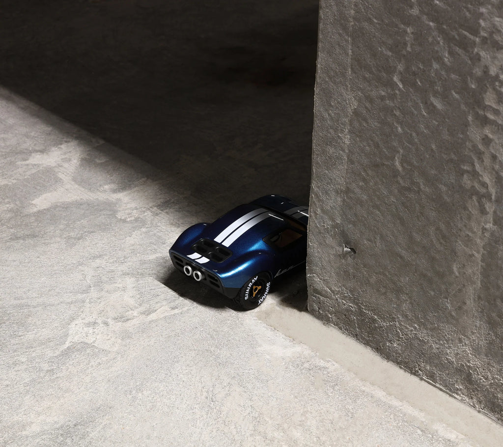 A Playforever MINI KEN CASH Blue car, inspired by 1960s endurance racing with white stripes, sits partially behind a textured concrete wall on a smooth, light gray surface.