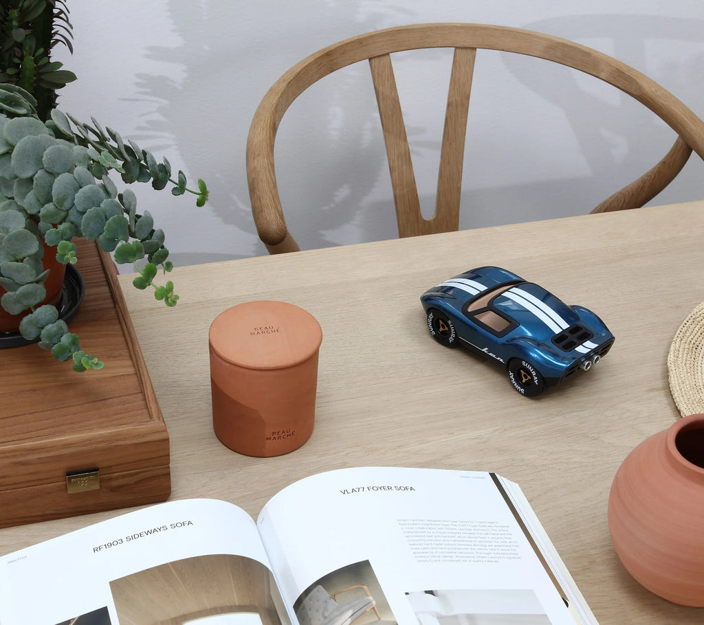 A wooden table featuring the Playforever MINI KEN CASH Blue racing car, inspired by 1960s endurance racing, a terracotta container, potted plant, open magazine, and a wooden chair in the background.