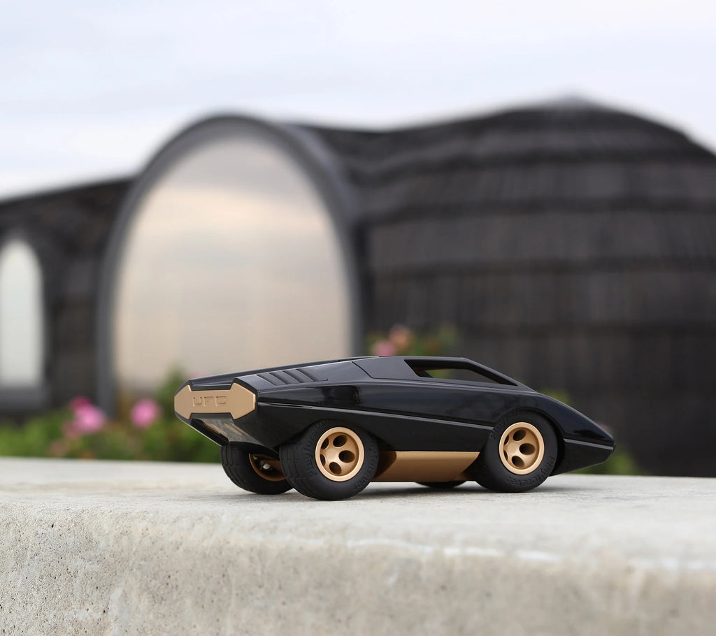 The Playforeer UFO Cannone Car in Black, featuring a sleek geometric design inspired by 70’s concept cars, sits on smooth concrete. A modern round building with a dark wood exterior is blurred in the background.