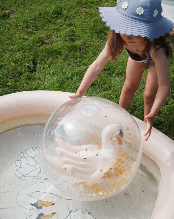 A young child in a swimsuit and sun hat is playing with a 40 cm Inflatable Swan Beach Ball filled with glitter and confetti in a small kiddie pool on a grassy area.