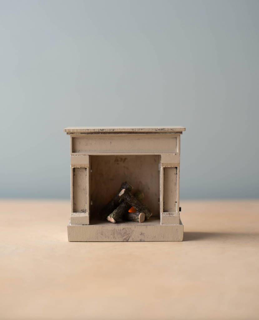 A small beige Maileg Cozy Christmas Set fireplace with faux logs sits on a light surface against a muted blue background—an ideal piece for your holiday collection.