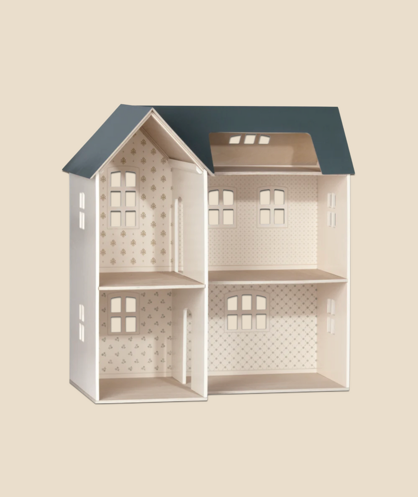 A Maileg dollhouse with an open back, revealing empty rooms with patterned walls, set against a neutral background. The house has a navy blue roof and a light beige exterior.