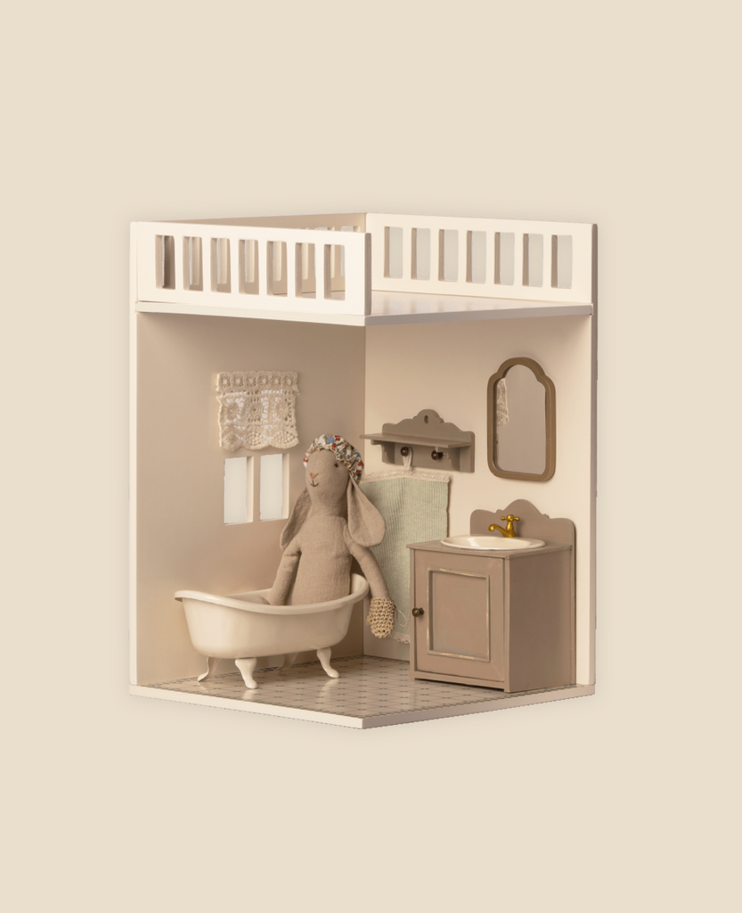 A minimalist Maileg House of Miniature Bonus Room - Bathroom in the dollhouse featuring a white bathtub, a wooden cabinet with a sink, a small mirror on the wall, and a plush doll in a hat and scarf. The color palette is neutral.