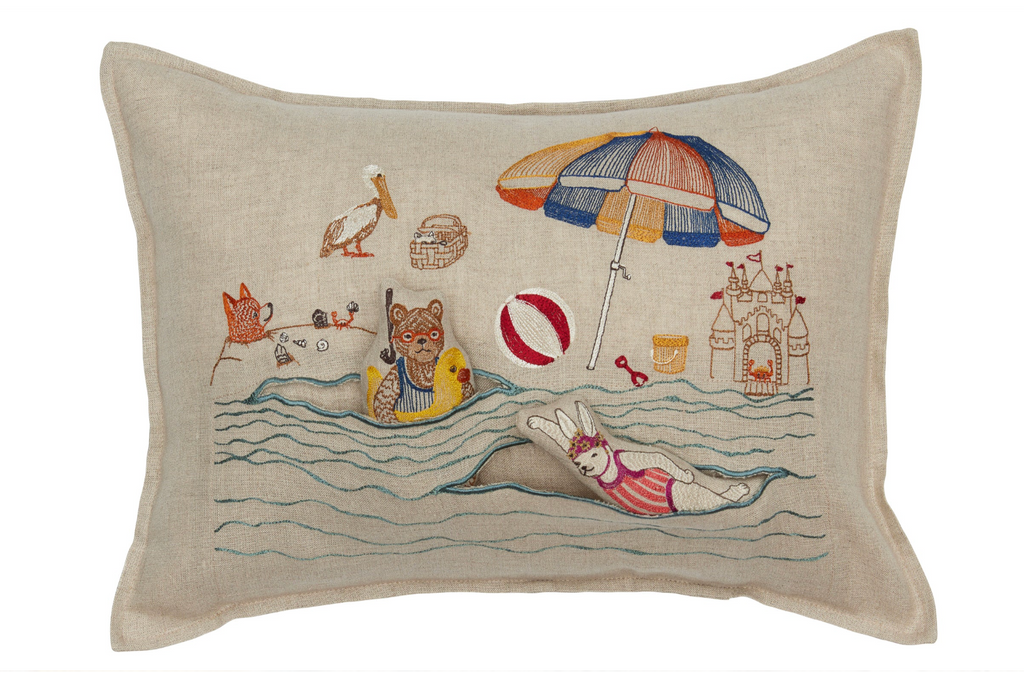 A decorative Coral & Tusk Day at the Beach Pocket Pillow with a beach scene embroidery featuring a bear and a rabbit floating on the ocean, surrounded by a beach umbrella, sandcastle, seagull, and beach.