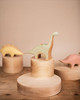 A handmade Wooden Brachiosaurus Dinosaur toy dinosaur stands on a circular wooden platform with other handcrafted wooden toys and a wooden figure partly visible in the background.