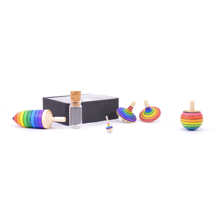 The Spinning Top Champion Learning Set - Rainbow features five vibrant tops in different sizes, displayed before a black box and glass corked bottle on a white background—ideal for skill testing and compliant with toy safety standards.