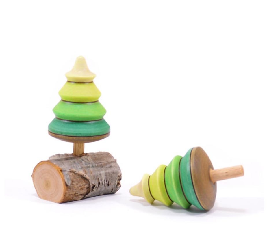 The Mader Spinning Tree Top On Trunk features two pine tree-shaped maple wood toys with stacked green discs—one upright on a log base, one lying on its side—both meeting strict toy safety standards against a white background.