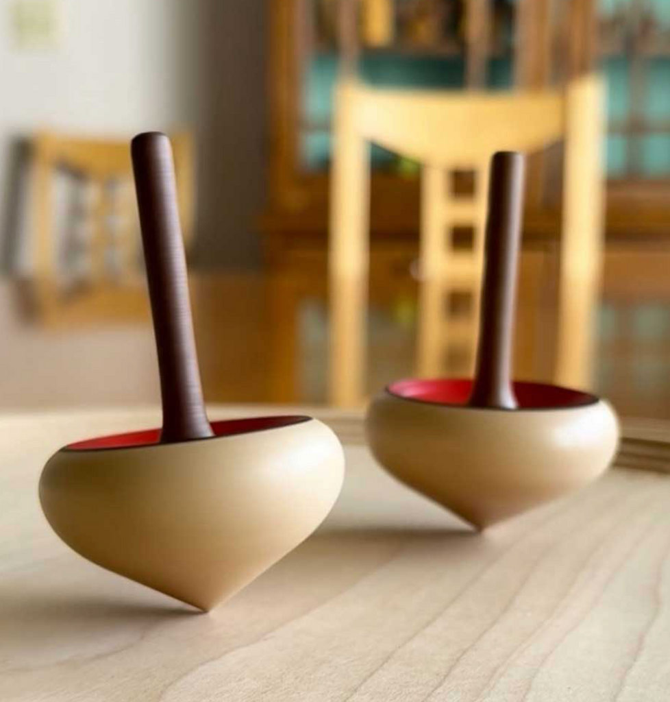 Two Mader Zwirbel Spinning Tops - Green, crafted from Walnut and Ash with dark handles and red interiors, rest on a smooth light-colored surface. A wooden chair and cabinet appear softly blurred in the background.