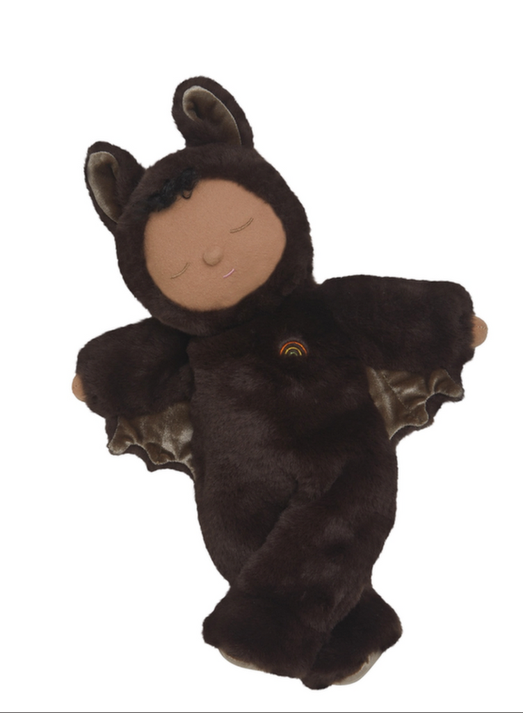 The Olli Ella Cozy Dinkum - Bat Ozzie - Dark Umber is a posable plush doll with a tan face, closed eyes, and a brown furry bat costume featuring bat ears, wing-like arms, and a small rainbow patch on its chest. Background is white.