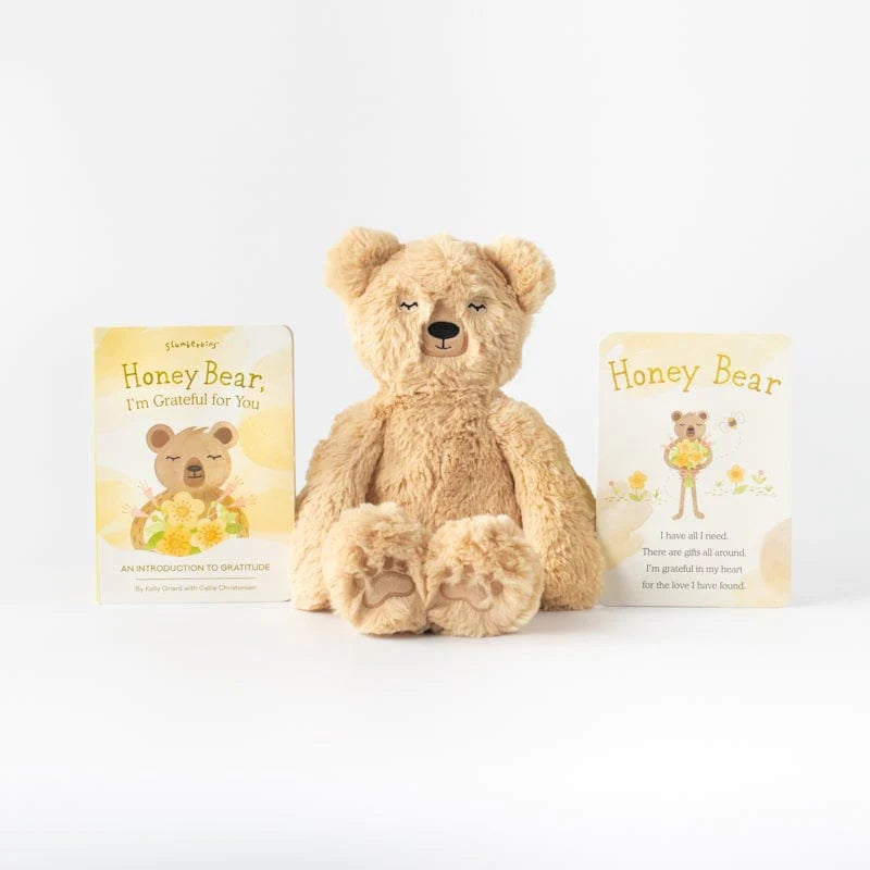A Slumberkins Honey Bear Kin + Lesson Book On Gratitude, named Honey Bear, sits centered between two children's books titled "honey bear, i'm grateful for you" on a plain white background.