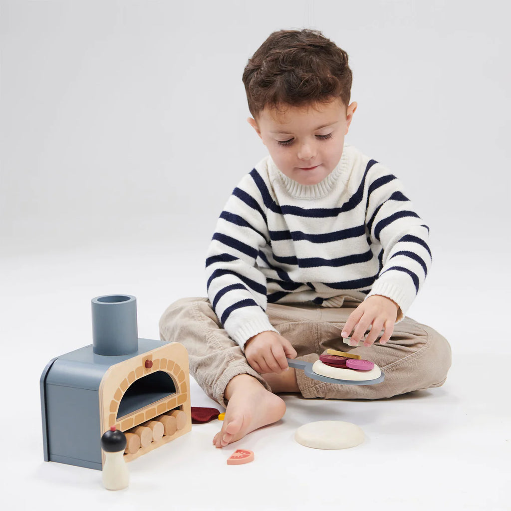 A young child in a striped sweater engages in pretend play with a Wooden Make Me A Pizza Set on a white background, slicing a toy eggplant with a toy knife.