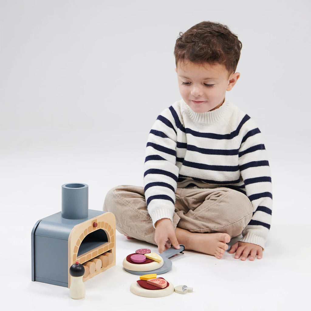 A young boy wearing a striped sweater and beige pants is engaged in pretend play with the Wooden Make Me A Pizza Set on a white background. He appears focused on cutting a toy pizza with various toppings.