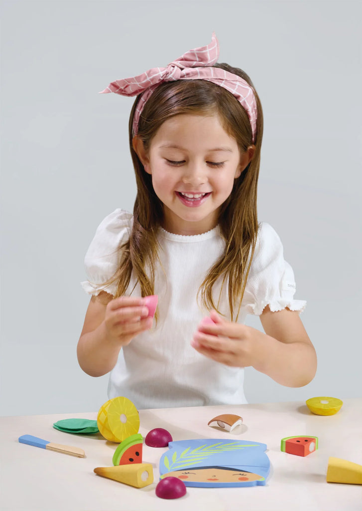 A young girl with a pink headband is smiling and playing with a Tropical Fruit Chopping Board on a table, each piece connected by velcro for easy cutting, holding a piece of toy fruit in her hands.