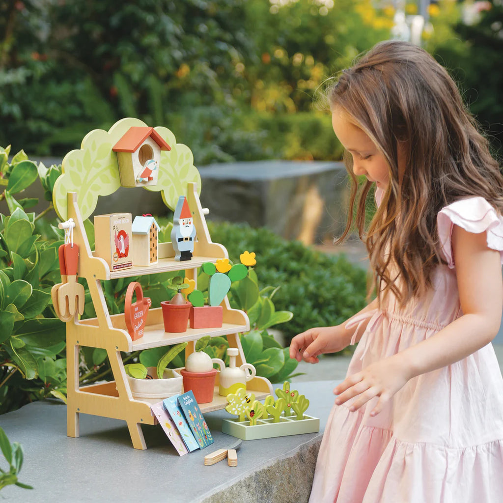 A young girl in a pink dress enjoys pretend play with her Garden Center outdoors, complete with small pots, plants, tools, and a birdhouse on a tiered shelf. Lush greenery and a stone path create the perfect backdrop for developing social and language skills.