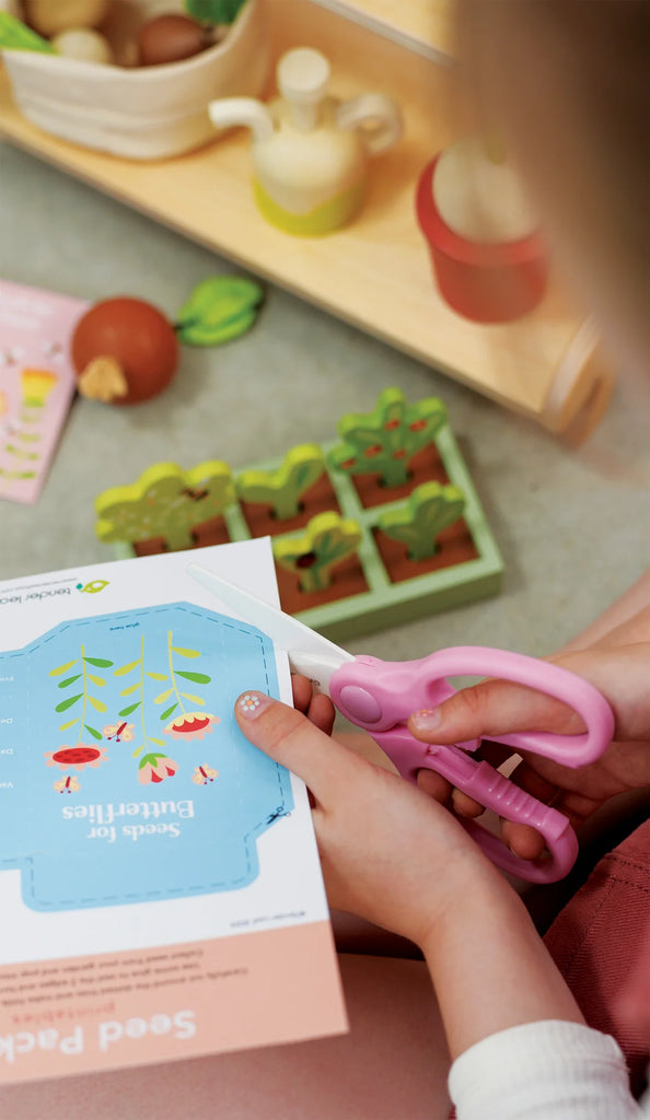 In the Garden Center, a child uses pink scissors to cut out "Seeds for Butterflies" paper with butterfly and plant designs. This imaginative play, featuring toy plant models and wooden vegetables, promotes social and language skills in a colorful environment.