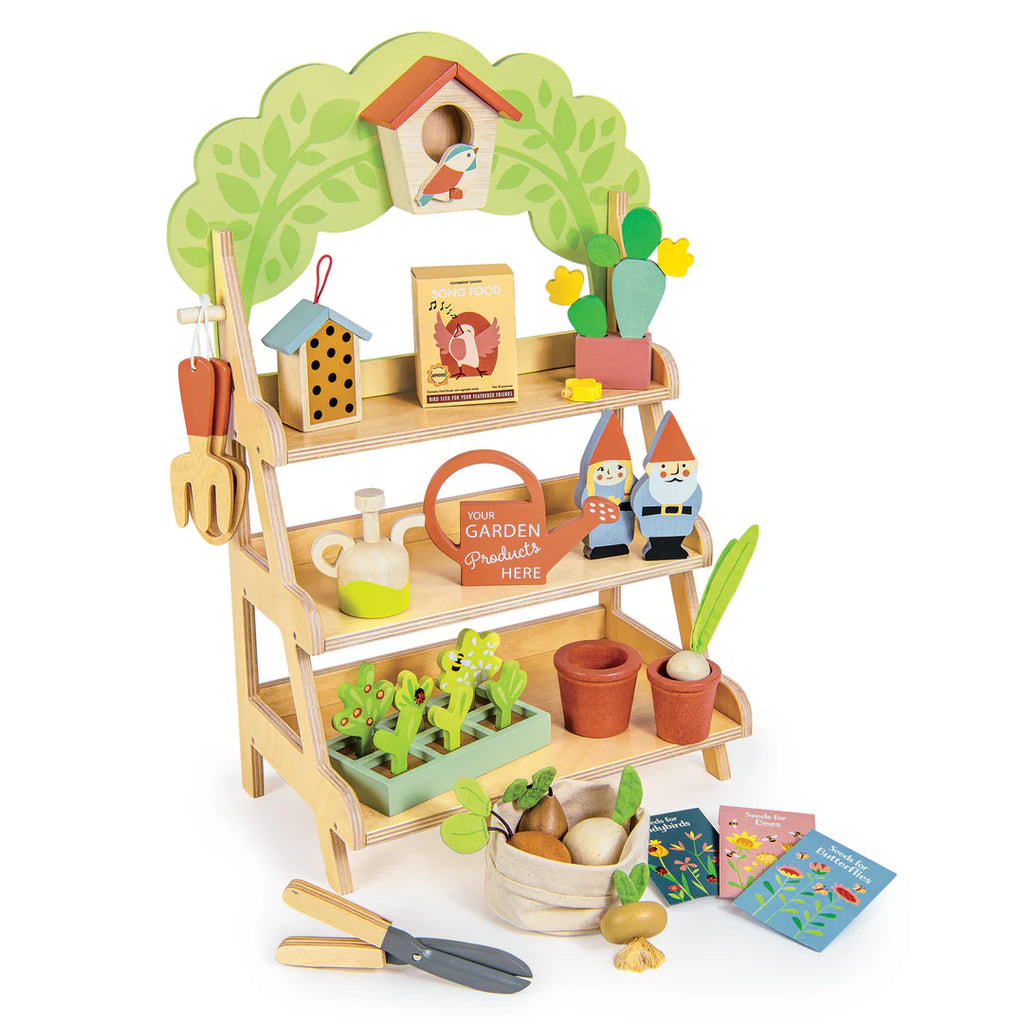 The Garden Center is a toy wooden stand for pretend play, featuring shelves with garden-themed accessories like 2 gnomes, potted plants, gardening tools, seed packages, and a basket of vegetables. It has a tree-shaped backdrop with a birdhouse to enhance the playful experience.