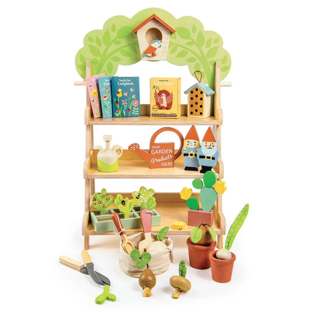 The Garden Center is a colorful wooden playset that mimics a bustling garden center, featuring shelves with a miniature birdhouse, books, gnomes, tools, plants, seed packets, and pots. It's perfect for pretend play under an arch formed by a tree and leaves.