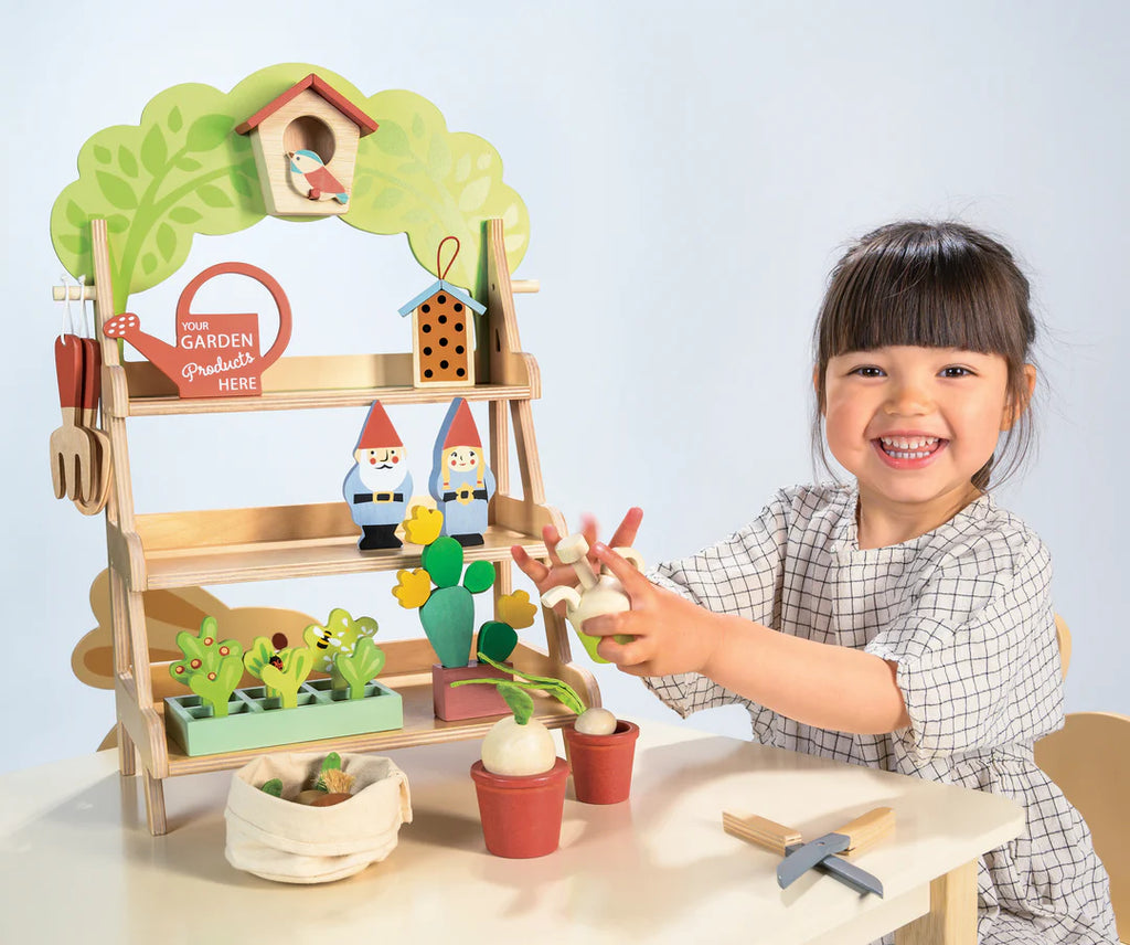 A smiling dark-haired child in a checkered dress enjoys pretend play with the Garden Center, a wooden toy set. It includes miniature gardening tools and potted plants on shelves, inviting kids to indulge in their own magical garden adventure.