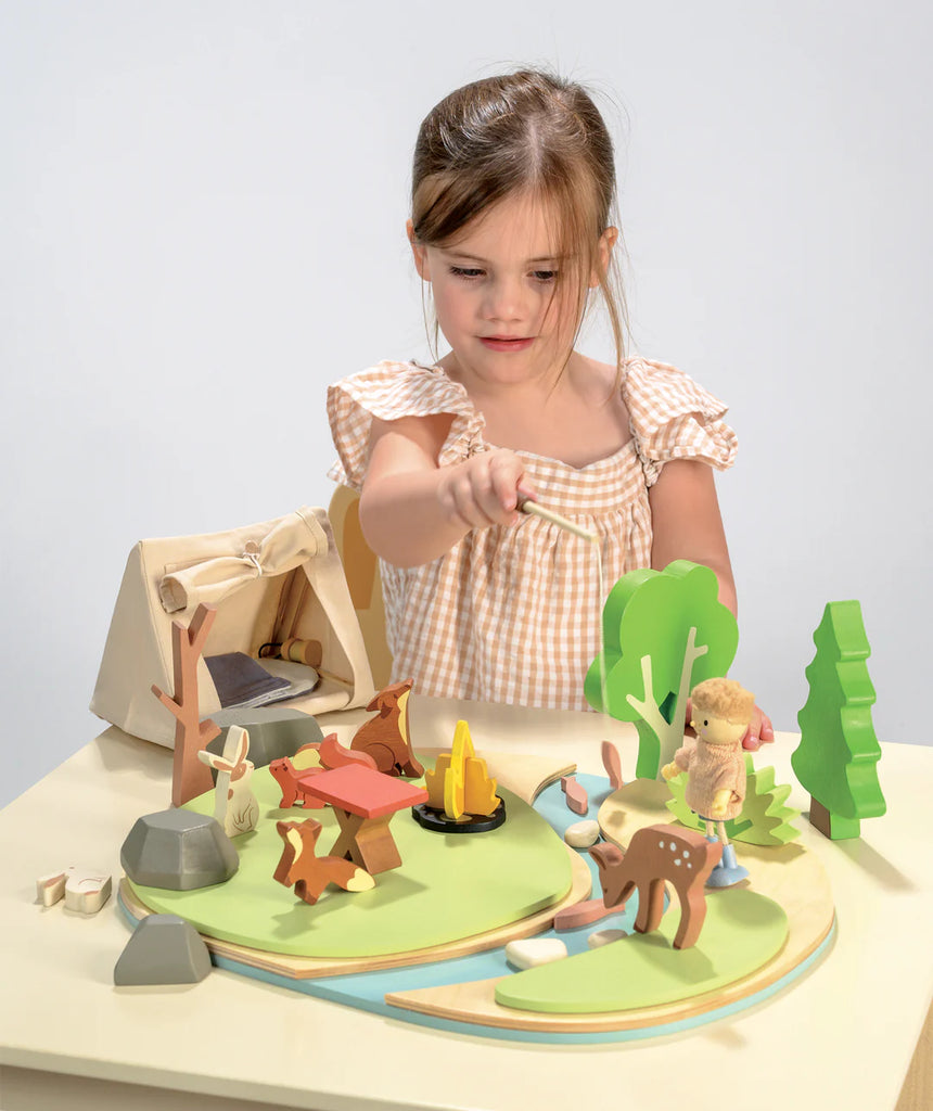A young girl in a checkered dress immerses herself in imaginative play with the Wildlife Camping Playset on the table. Featuring trees, animals, a campfire, tent, and stream, she skillfully weaves storytelling into her interactions with this captivating set.