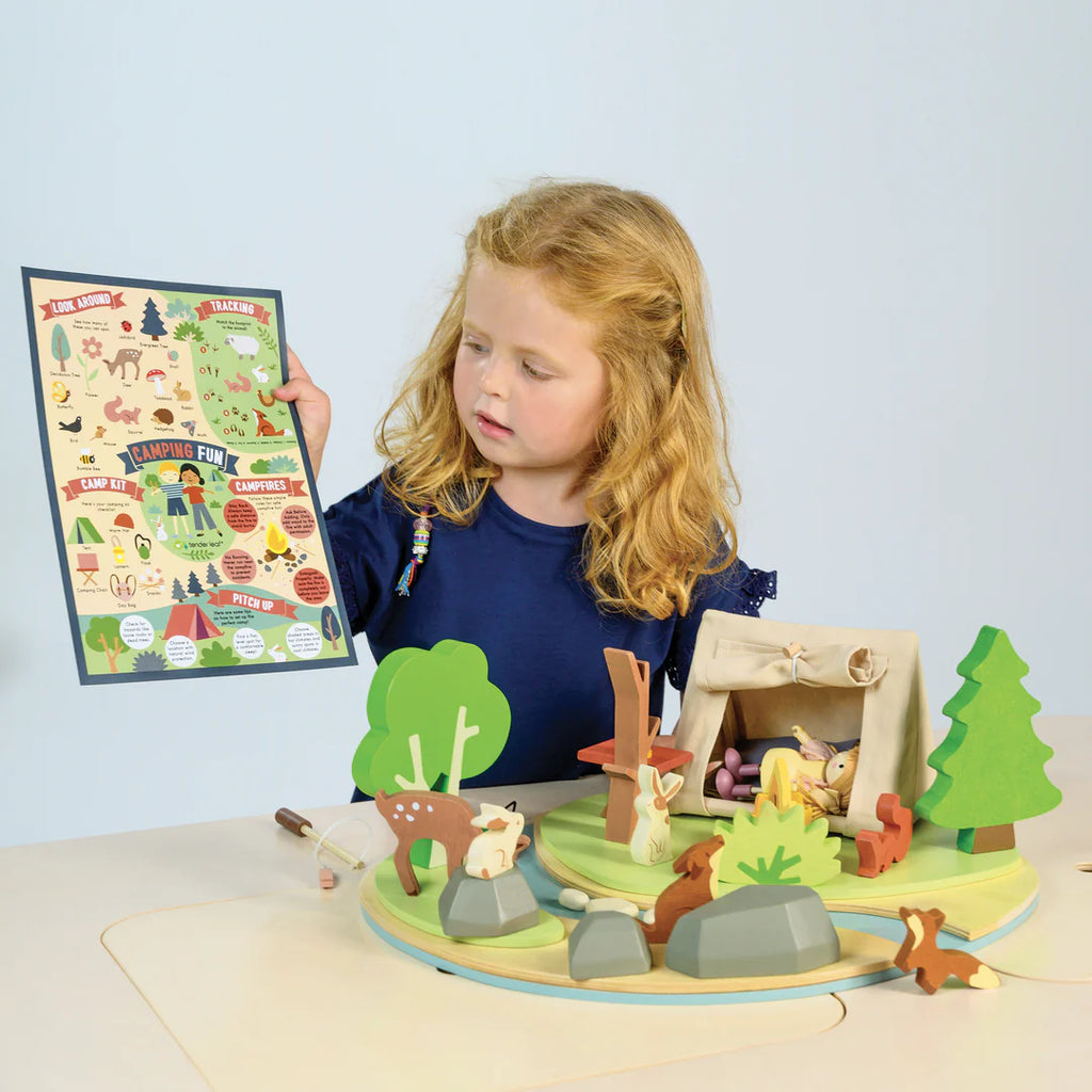 A child with long hair in a blue shirt holds a colorful camping-themed poster. In front is the Wildlife Camping Playset, perfect for imaginative play, featuring a tent, trees, rocks, and animal figures on a circular base. The background is light blue.
