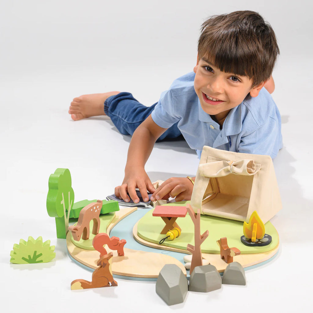 A young boy in a blue shirt plays imaginatively on a white surface with his Wildlife Camping Playset. He smiles while lying down, using the tent, campfire, animals, and trees to craft stories that bring this miniature world to life.