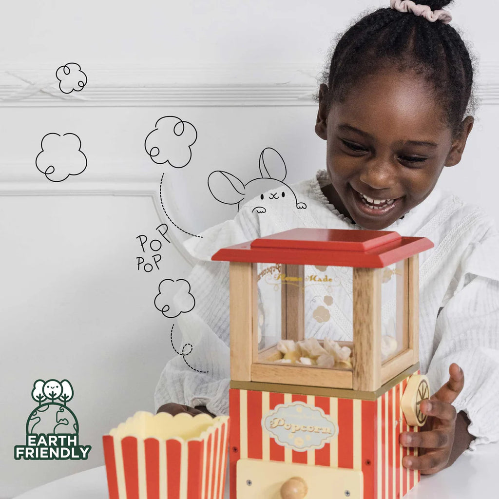 Young girl smiling while playing with a Vintage Popcorn Maker made of sustainable wood. Next to the machine is a container of popcorn. Hand-drawn doodles of a bunny and popcorn popping are on the wall behind her, encouraging fine motor skills development. The "Earth Friendly" logo is in the bottom left corner.