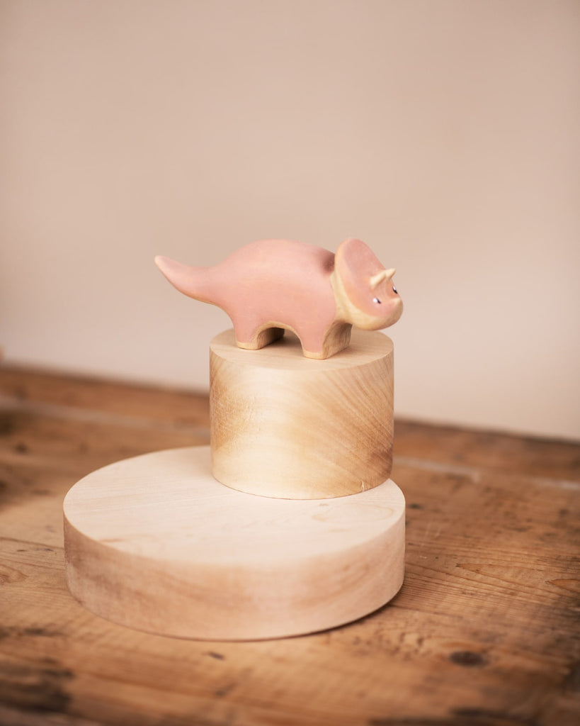 A pink ceramic Handmade Wooden Triceratops Dinosaur figurine, coated with non-toxic paint, stands atop two stacked wooden cylinders, placed on a wooden surface against a soft beige background.