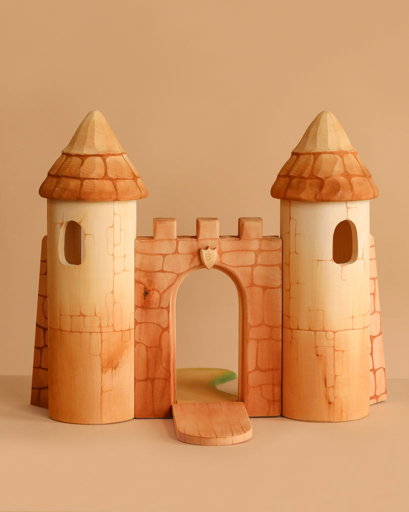 The Bumbu Handmade Wooden Castle features two round towers, an arched entrance, and a drawbridge. Crafted from smooth, light-colored wood with painted brick details, it's perfect for small world play and displayed against a plain beige background.