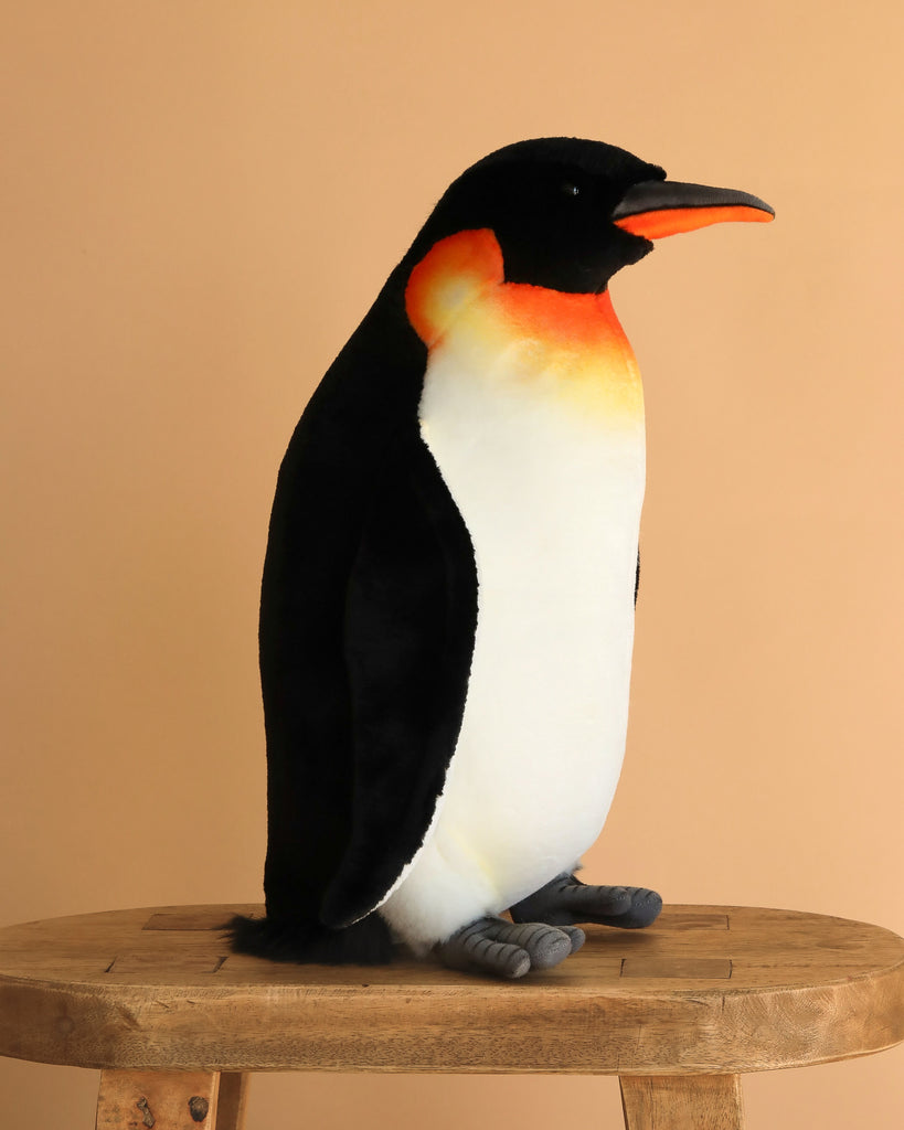 The Penguin Stuffed Animal, Medium, features lifelike black, white, and orange details and sits upright on a wooden stool against a beige background, showcasing the realistic design found in HANSA animals and similar plush toys.