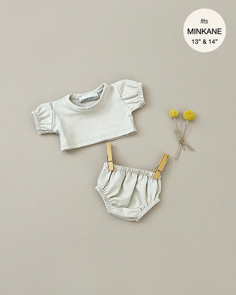 A light grey doll outfit, part of the Les P'tits Basics collection, is displayed against a beige background. The set includes a balloon-sleeved t-shirt and elasticated shorts. Wooden clothespins clip the shorts to a string, while two yellow billy button flowers lay beside the outfit. Text reads "Minikane Doll Clothing | Charlotte Balloon Sleeve T-Shirt and Briefs Set in Linen Ecru fits Minikane Gordis dolls 13' & 14'".