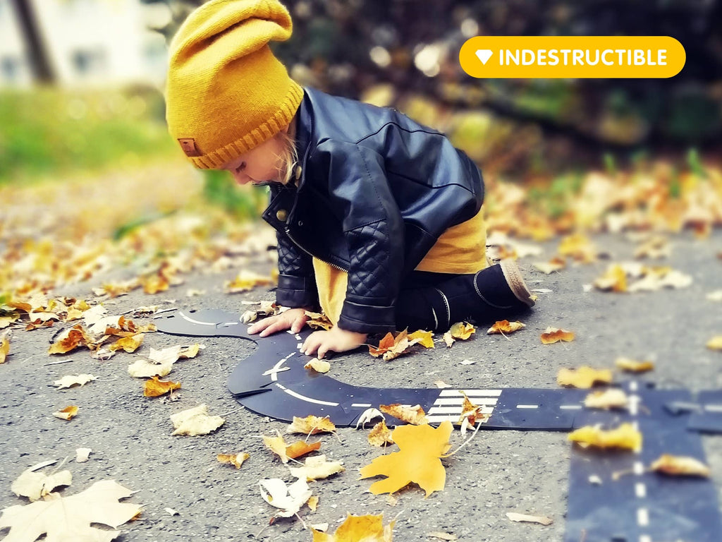 A young child in a yellow hat, yellow dress, and black leather jacket kneels on a leaf-covered pavement, playing with the WaytoPlay: Extra Long Flexible Road Set - King of the Road. A label reading "INDESTRUCTIBLE" appears in the top right corner.