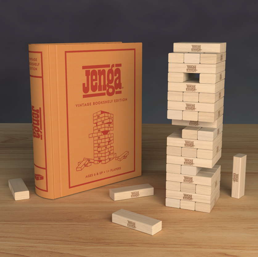 The Jenga Vintage Bookshelf Edition, styled like a book, stands upright by a partially played tower and loose wooden blocks—ideal for family game night on a wooden table.