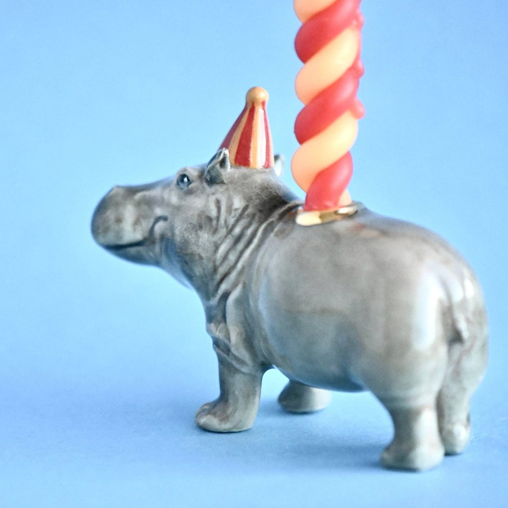 A small figurine of a Hippo Cake Topper, hand painted and crafted from fine porcelain, with a colorful twisted candle attached to its back, set against a light blue background.