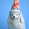 A close-up of a small fine Poodle Cake Topper figurine of a smiling white bear wearing a striped party hat against a soft blue background.