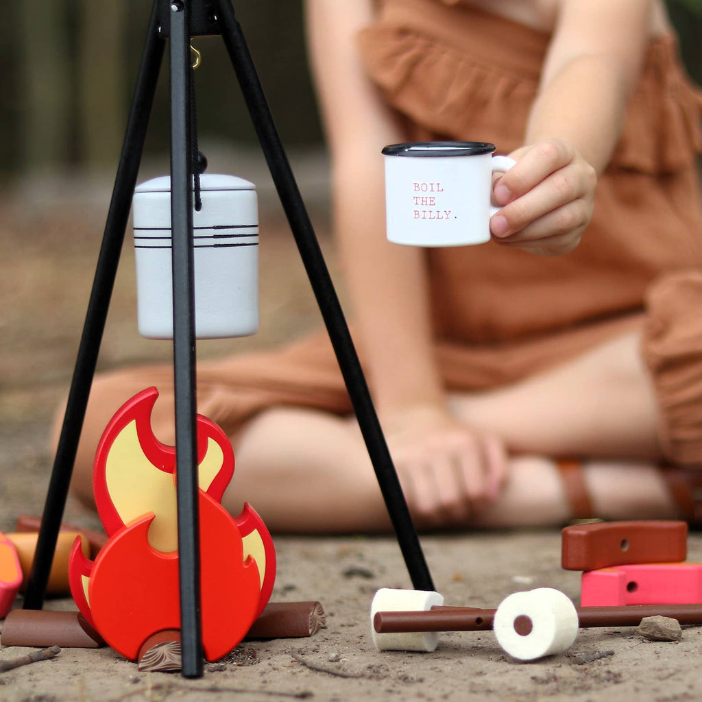 A child in brown clothes sits on the ground, holding a small white mug near the Wooden Camp Fire Set, which features a hanging pot on a tripod and scattered wooden food toys for kids’ pretend play.