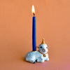 A Goat Cake Topper shaped like a smiling lamb, featuring a lit birthday candle, set against a soft peach background. The lamb is adorned with a party hat and a decorative bell.