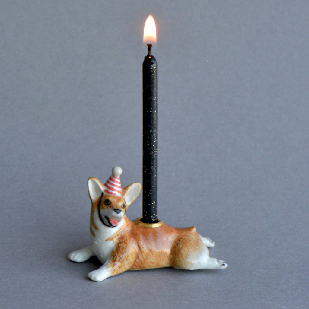 A whimsical hand-painted porcelain Corgi Cake Topper featuring a figurine of a smiling corgi wearing a party hat, supporting a lit black taper candle on a gray background.