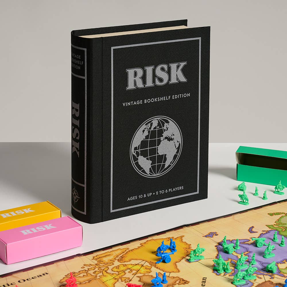 The Board Game: Risk Vintage Bookshelf Edition features a black, book-shaped box standing upright beside a 1996-style game board, colorful pieces, RISK cards, pastel card boxes, and small army figures atop a world map.