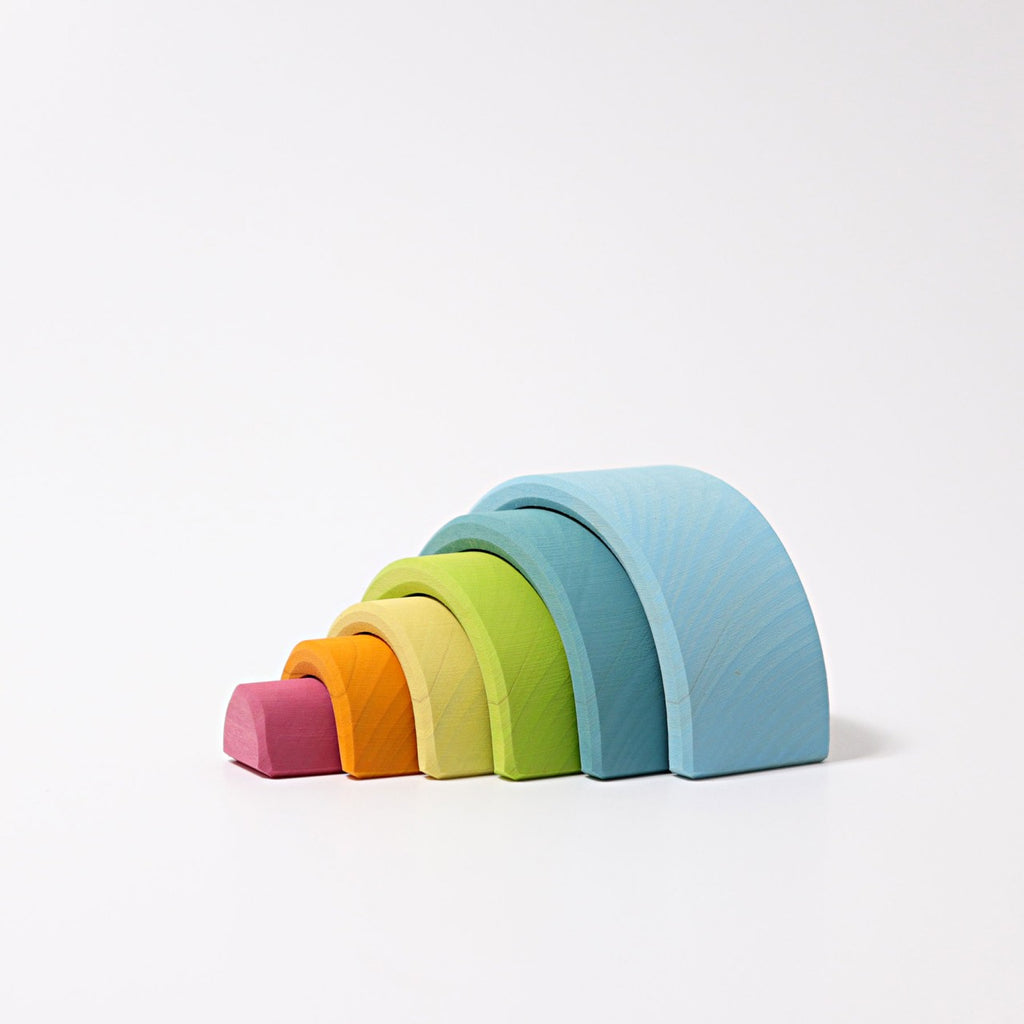A versatile toy featuring a set of colorful wooden rainbow stacking arches arranged in a gradient. Crafted from lime wood, the smallest arch is red, followed by orange, yellow, light green, dark green, and the largest arch is blue. The arches are stacked neatly from smallest to largest on a white background. 
Introducing Grimm's Small Wooden Rainbow, Pastel!