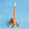 A lit orange spiral candle placed on a small, hand-painted Octopus Cake Topper against a pale blue background.