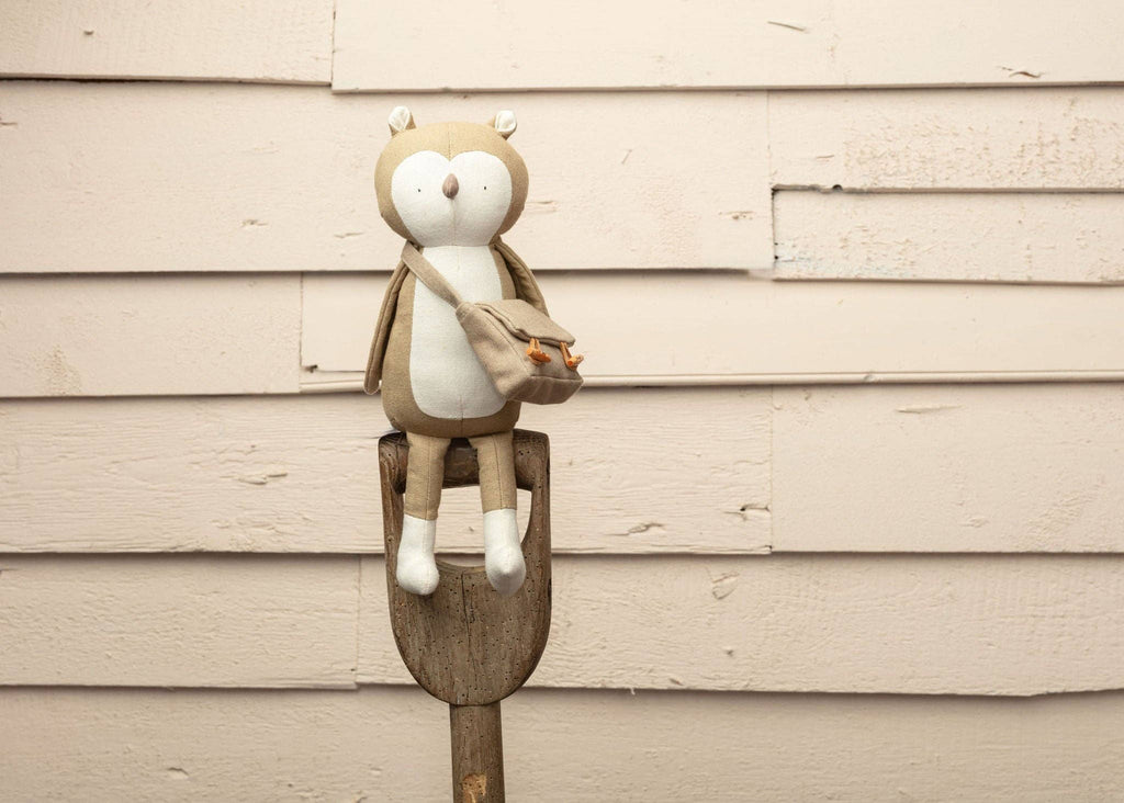 Owen The Owl, a plush toy featuring a messenger bag, nicely perches on the handle of a shovel. It adds charm to a woodland-themed scene against a wooden wall with peeling paint.