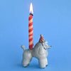 A Poodle Cake Topper shaped like a poodle, with a red and white twisted candle lit on its back against a plain blue background.