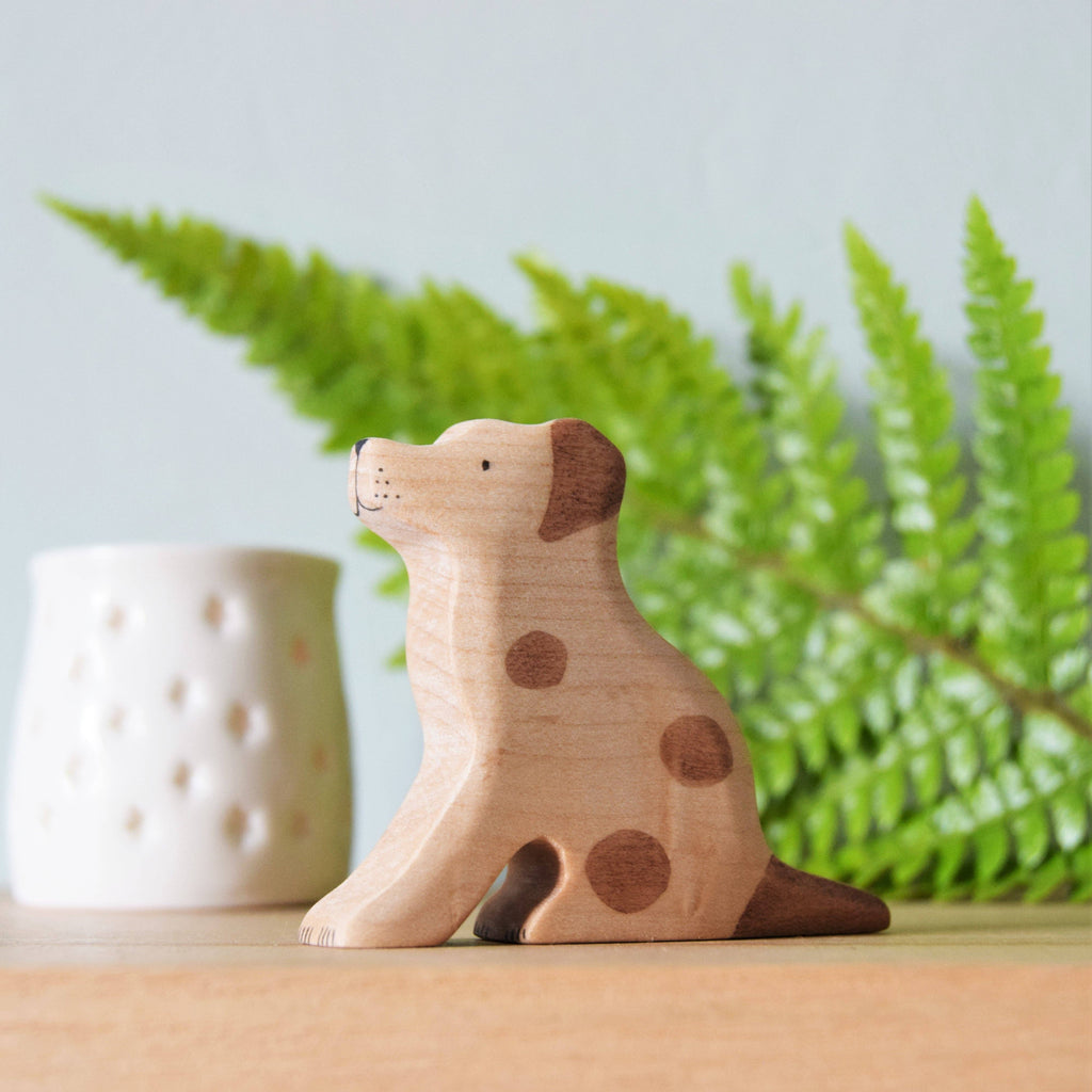 A Handmade Wooden Jack Russell toy rests on a wooden surface, adorned with brown spots from natural plant-based stains. Behind it, a green fern leaf and a white perforated ceramic pot enhance the scene against a soothing light blue backdrop.