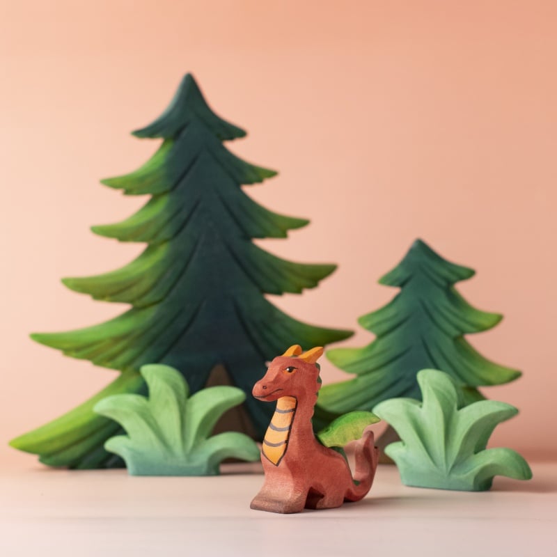 The Bumbu Handmade Wooden Baby Dragon features yellow accents and stands before stylized green trees on a soft peach background—a charming Montessori wooden toy perfect for imaginative play.