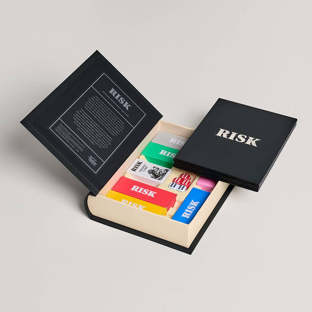 The Board Game: Risk Vintage Bookshelf Edition features a stylish, book-shaped box with vintage design that opens to reveal colorful RISK cards, game pieces, and dice all neatly organized inside.